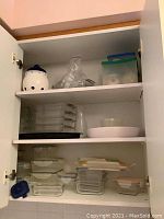 Photo of kitchen cabinet shelves showing four glass loaf pans, multiple glass food storage containers with lids, plastic containers with colored lids, white ceramic bowls, and a ceramic container with holes and blue lid.