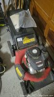 Front angled view of the red and black Yard Machines lawn mower, showing engine top, wheels, and handlebar with manual in plastic