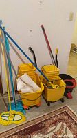 Photo showing three yellow mop buckets, mop handles and mop heads leaning against a white wall and a carpeted floor.