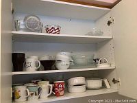 Photo of cupboard shelves showing a variety of ceramic mugs, clear glass bowls, white ceramic bakeware, and other kitchenware.