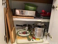 Photo showing interior of kitchen cabinet with various kitchen items including wooden cutting boards, metal and glass bakeware, a green salad spinner, and a Lagostina stainless steel pot steamer.
