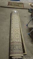 Rolled beige area rug with floral pattern and dark blue border on a concrete floor.