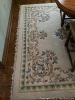 Full view of wool area rug showing floral border design and central floral motifs, placed next to wooden table and chair. The rug shows signs of wear and light fraying at edges.