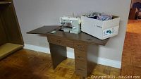 Side view of wooden sewing cabinet with fold-out work surface, PFAFF 362 Automatic sewing machine installed and cardboard box of sewing notions on top.