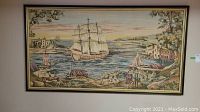 Full view of framed tapestry wall hanging showing sailing ship, shore buildings, trees, people, and ocean scene