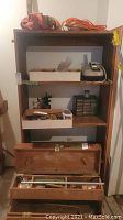 Full view of shelving unit with toolbox open on bottom shelf, white trays with tools and hardware on middle shelf, and phone and organizers on top shelf. Red and orange straps on top of shelving unit.