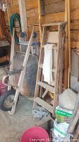 Wooden step ladder with cloth draped over steps, wooden boards and poles behind, metal wheelbarrow beside it, garden products on floor