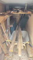 Photo showing wooden lumber boards, rolled green wire fencing, metal wheelbarrow, and scaffolding parts stacked in a narrow space.