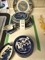 Overall view of a stack of blue and white plates with a small creamer and a decorative tray, together with an Aruba souvenir plate in the background. The dishes feature Willow pattern designs.
