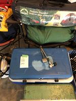 Photo showing green tent bag labeled Escort Pentagon Dome Tent, blue two-burner camp stove with some tools on top, blue and white cooler beneath stove.
