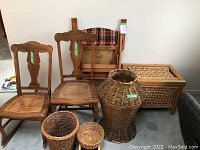 Photo showing two wooden chairs with carved backs, one with a woven seat, the other with a wood seat; various woven baskets including a tall vase-shaped basket, smaller round baskets and a rectangular woven storage bin with inscription.