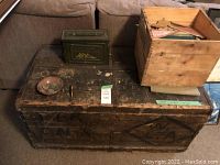Wooden chest with stenciled designs, green military ammo box, and small wooden crate on top, placed in front of a brown couch.