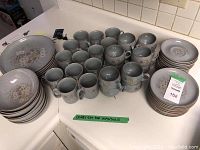Overview of entire Denby stoneware dinnerware set showing stacked dinner plates, bowls, mugs, teacups, and saucers arranged on countertop, labeled for Maxsold.