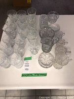 Full lot of glass and crystal items displayed on a white table, including multiple high ball glasses, wine glasses, creamers, sugar bowls, and small dishes with decorative cut and etched patterns.