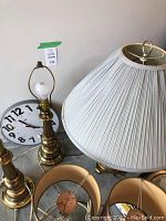 Two tall brass lamps without shades and a round white wall clock with black numbers on the floor next to a tall brass lamp with beige pleated lampshade.