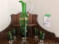 Full view of green glass decanter and five matching glasses arranged on wooden table with wood backdrop. Shows gold fleur de lis pattern and wear on gold detailing.