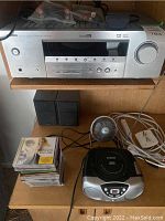 Photo showing Yamaha RX-V470 receiver on top shelf, Sylvania CD player and radio on bottom shelf, small fan, stack of CDs, and power strip cords.