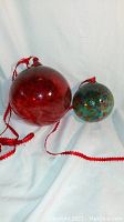 Photo showing two glass balls, one larger red and one smaller multicolored, both with attached red ribbons for hanging.