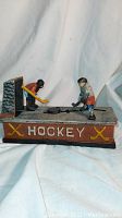 Front view of the cast iron hockey mechanical bank showing two hockey players facing each other over a puck, with the word 'HOCKEY' and crossed sticks painted on the side.