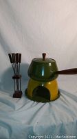 Vintage fondue set with green pot on yellow burner base and six metal serving forks with wooden handles in wooden stand.