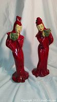 Full view of two tall red chalkware figurines dressed in theatrical costumes holding green masks.