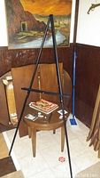 Black wooden artist easel shown standing on tiled floor against corner wall, angled to display front and one side, with adjustable support beam visible.
