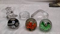 Image showing six glass and crystal paperweights on white fabric: clear glass elephant, rectangular floral paperweight, two spherical floral paperweights, black and white Pokemon Pokeball with Charmander, and Mats Jonasson dolphin figurine.