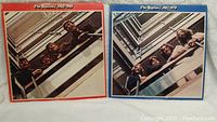 Photo showing front covers of two Beatles albums, one with red border titled '1962-1966' and one with blue border titled '1967-1970'. Both feature the band members looking upwards on a stairwell.