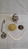 Photo showing all items: one brass ornate Western style belt buckle, two vintage necklaces (clear beaded and silver-tone with floral pendant), a decorated floral egg trinket that opens, and two small decorative trinket boxes (cream and black).