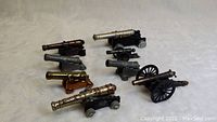 Photo showing all eight small antique metal and diecast cannon replicas arranged on a white surface, highlighting diverse styles and finishes.