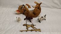Photo showing all seven dog-themed items arranged on a white surface: reclining chihuahua wine holder, three vintage dog ceramics, reddish-brown small dog figurine, RCA Victor dog salt shaker, brass hunting dog key holder shaped like a dog on a rifle.