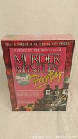 Front view of the boxed Murder Mystery Party game showing the title and theme, red and black design with graphics illustrating a murder mystery setting on a dancefloor.