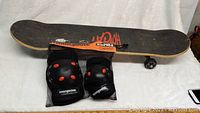 Photo showing the skateboard with grip tape and red logo, with the knee and elbow pads in mesh bag resting on the board.