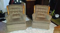 Two brown fabric swivel glider chairs with button-tufted backs and wooden framed armrests shown from the front on a carpeted floor.