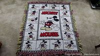 Top view of woven Mickey Mouse throw blanket laid flat on carpet, showing front side with multi-character design and colored fringe edges.