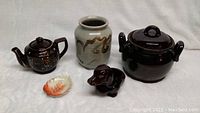 Collection of vintage made in Japan ceramic items including a teapot, food pot, vase, leaf dish, and dachshund figurine shown on white fabric background.