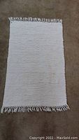 Full view of white hand woven floor mat with fringe laid flat on carpeted floor. Minor stains visible.