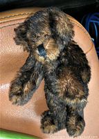Front and side views of a small chocolate brown teddy bear with glass eyes and black stitched nose sitting on brown leather. Fur appears dense and soft with lighter brown accents. Shows excellent condition with no visible damage.
