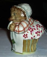 Side profile showing Mrs. Tiggy-Winkle figurine wearing bonnet and apron over red-checked blouse with floral patterned skirt and basket base