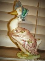 Front angled view of Jemima Puddle-Duck figurine wearing a blue bonnet and purple shawl with floral design, standing on green base.