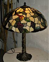 Front view of lamp showing illuminated stained glass shade with grapes and leaves design and detailed metal base.