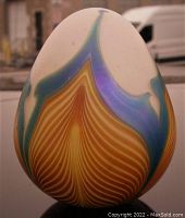 Front view of the iridescent Skookum egg paperweight showing the multicolored design with amber, purple, and teal patterns against a frosted white base.