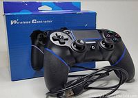 Black wireless PS4 controller with blue accents in front of its retail packaging box. Includes USB charging cable coiled beside it.