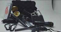 Photo showing emergency survival kit contents including compass, bracelets, whistle, clips, knife, fire starter, multi-tool, pen, flashlight in black protective case with foam padding.
