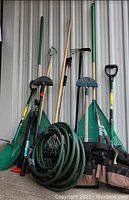 Photo showing collection of gardening tools including various shovels, rakes, hoes, a green coiled garden hose, and a brown and black tool belt with multiple pockets.