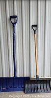 Photo showing two snow shovels leaning against a corrugated metal wall. One large blue plastic shovel with a wide scoop and push handle on the left. One smaller black plastic shovel with traditional wooden handle and D grip on the right.