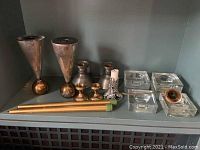 Shelf with two large metal conical candle holders with round gold bases, two smaller metal candle holders with round bases, a decorative silver-tone candle sleeve, two gold taper candles, and a white taper candle in a holder.