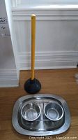 Photo showing stainless steel cat food and water bowls on a platter next to a yellow-handled black rubber plunger on wooden floor.