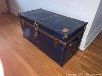 Full angled view of the blue chest showing the metal covering, locks, and general condition with visible wear and scratches on a wooden floor.
