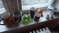 Overview of kitchen items on window sill including ceramic pitcher, stacked tea cups, clear storage jars, green glass covered dish, and small pepper grinder.